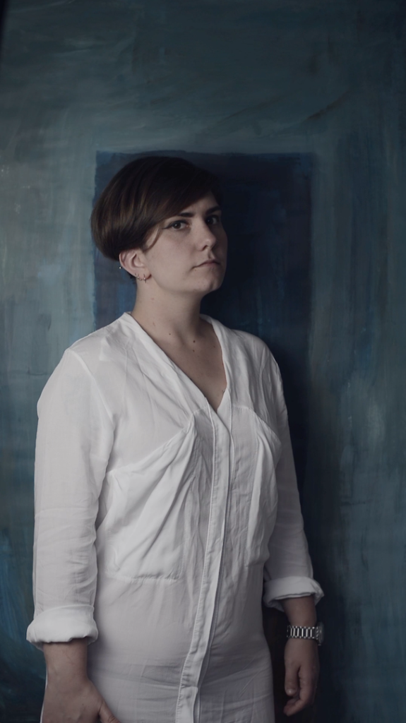 The artist in a white shirt on a blue painted background, looking at the viewer. 