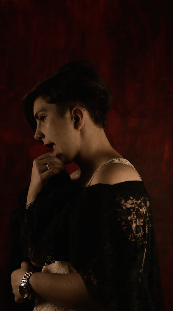 The artist viewed from the left side, wearing black lace and wiping her lip on her fingers, on a red background. 