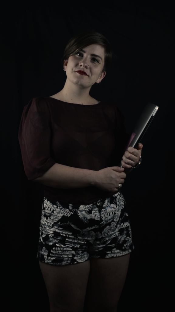 The artist smiling and making eye contact while holding her laptop on a dark background. 