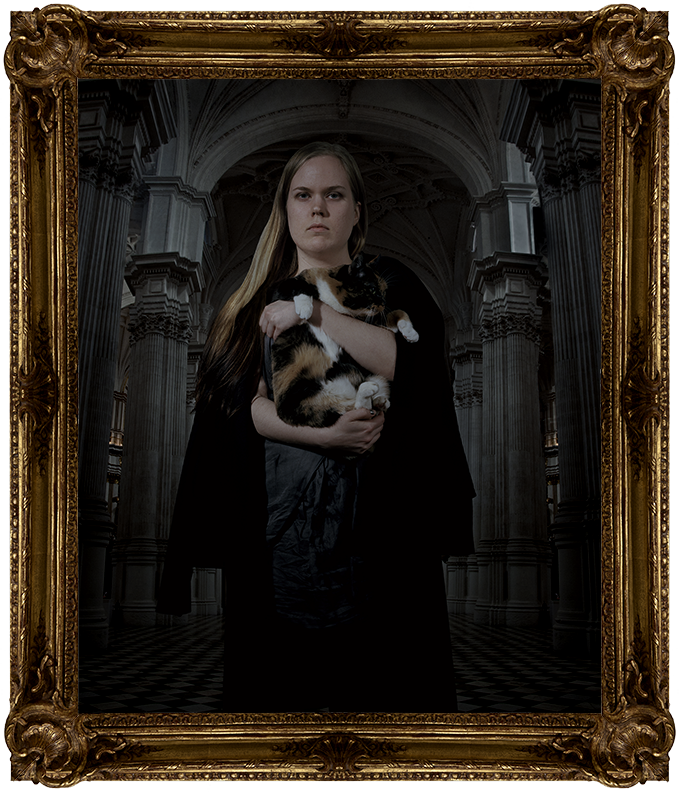 A woman wearing a black cloak, holding a cat. She stares sternly at the camera.