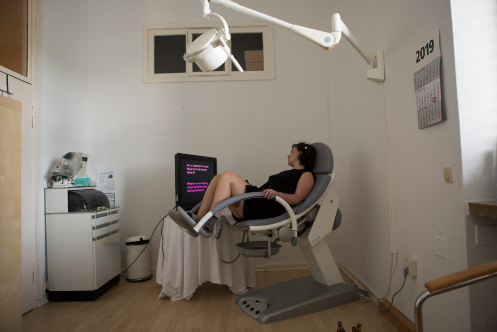 A person sitting in a gynecologist's chair and looking at the video artwork playing on screen.