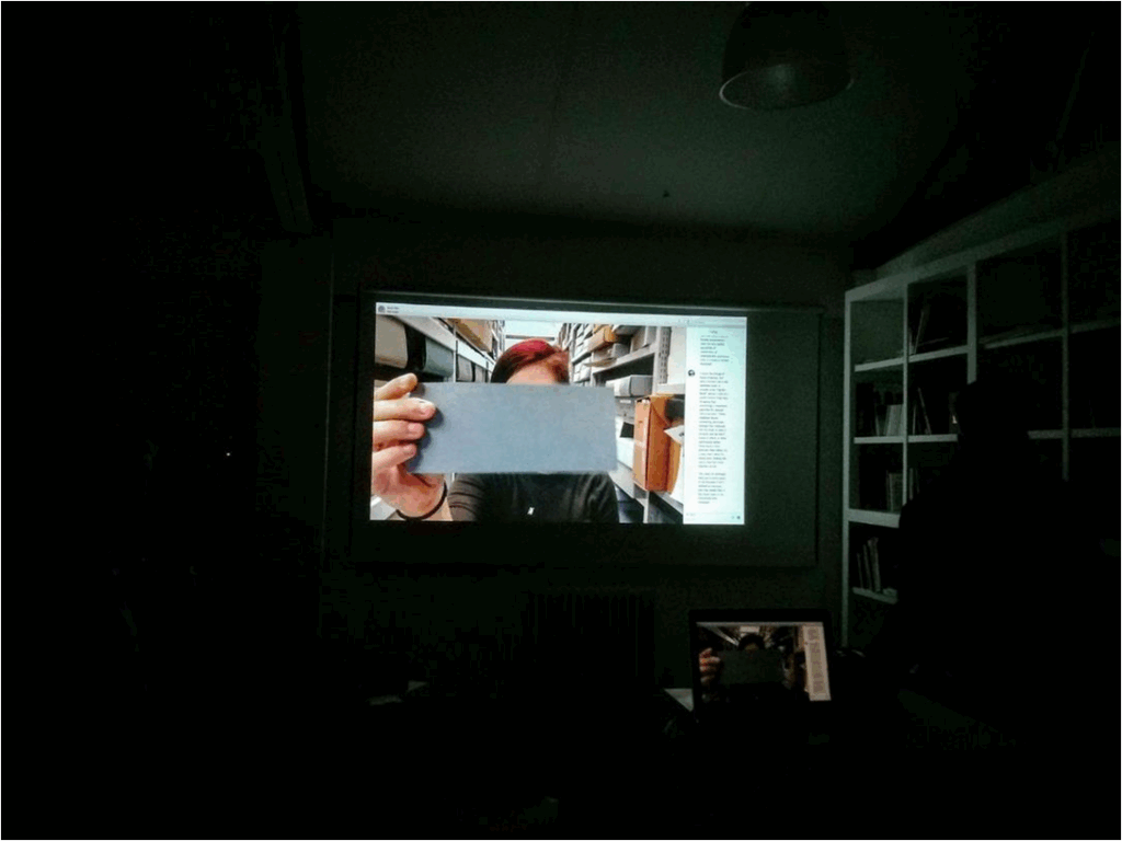 A projection of someone on a skype call in a dark room. The person is holding an item up to the screen. The item is a grey piece of paper.